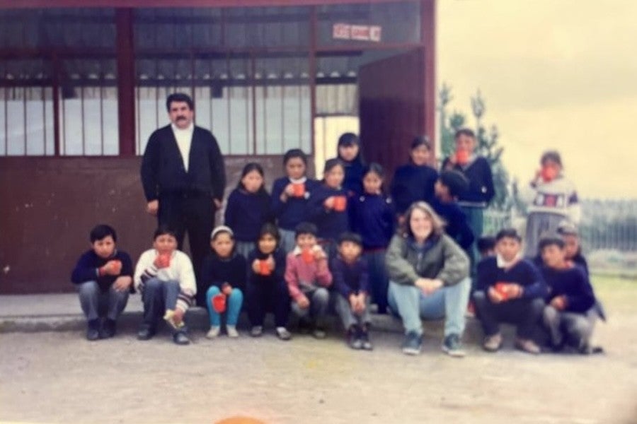 A male teacher and a class of small children sit with a woman in front of a school building.