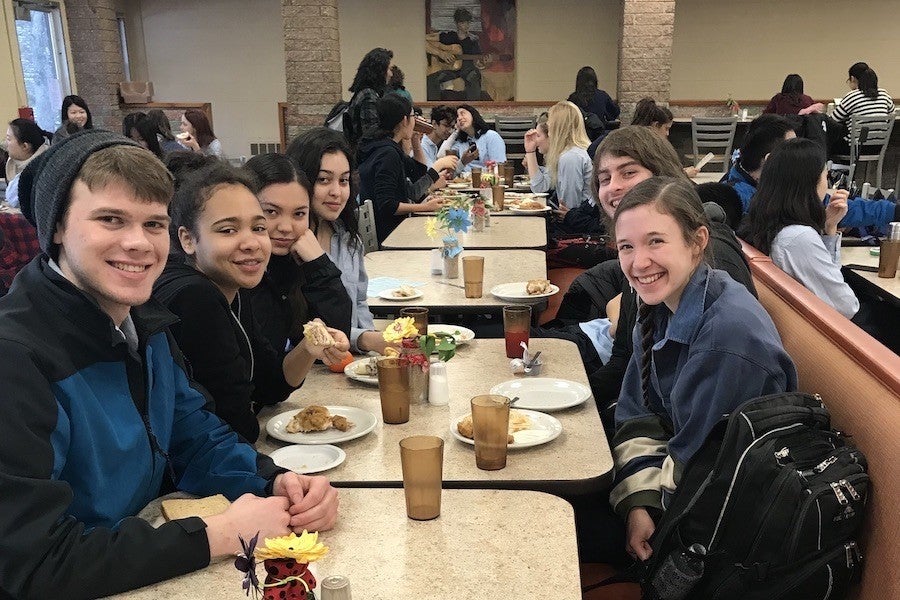 Six students sit around a table with plates, cups, and food. They smile at the camera. 