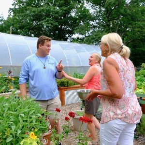 An instructor works with two adult students in a garden with a greenhouse in the background.
