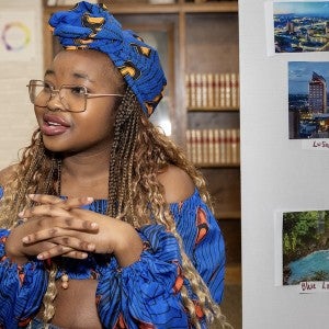 A student dressed in bright blue traditional Zambian garb presents from a poster board about her country. 