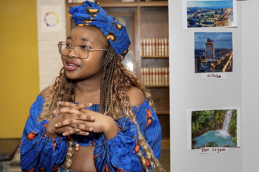 A student dressed in bright blue traditional Zambian garb presents from a poster board about her country. 