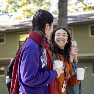 Smiling students stroll on campus with coffees in their hands.