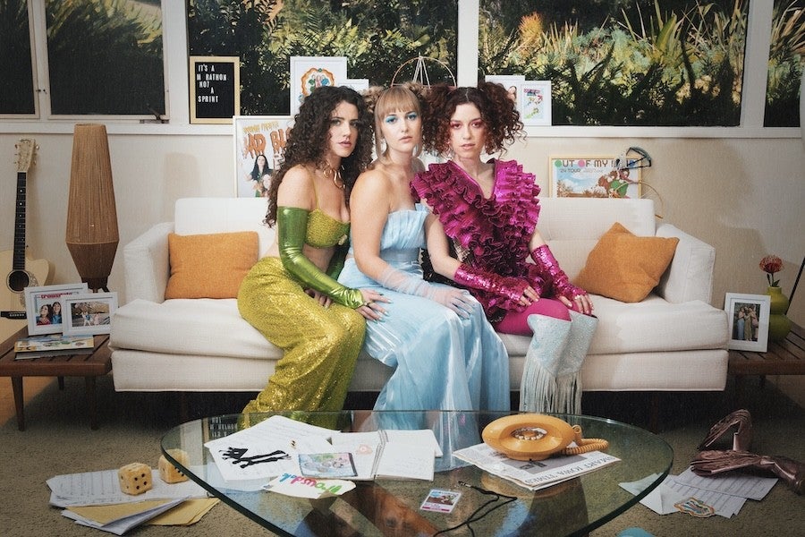 Three musicians dressed in vibrant colors sit closely on a couch in a messy room.