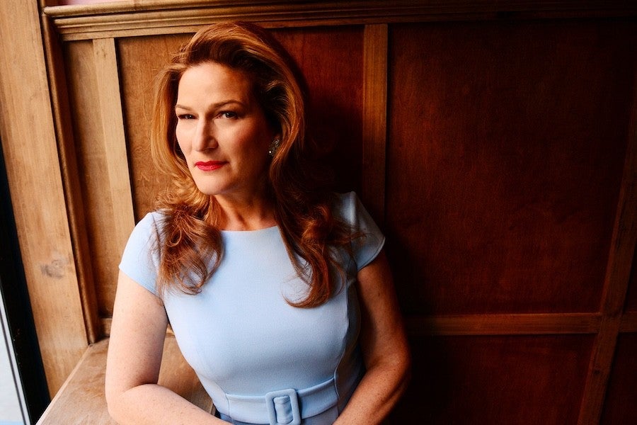 Ana Gasteyer seated beside a wooden paneled wall wearing a light blue dress.