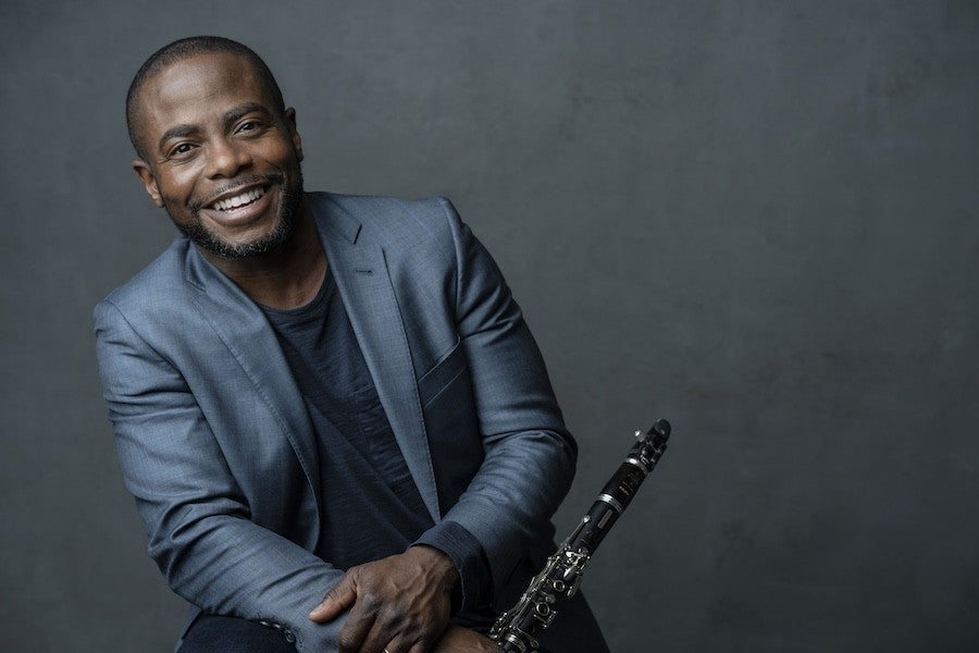 Anthony McGill stands in front of a grey background and smiles while holding his clarinet. 