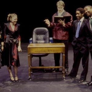 A theater performance with two women and three men on stage, standing around a wooden desk. A tall man in a lounge jacket jacket is reading a book books, while the two men on the right smile and address the two women on the left. 