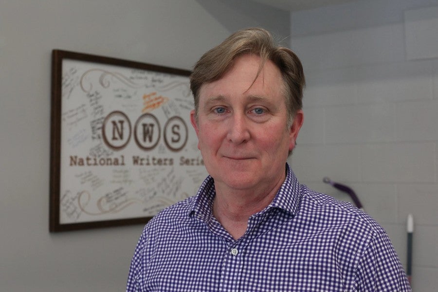 Doug Stanton smiles standing in front of a framed poster for the National Writers Series.