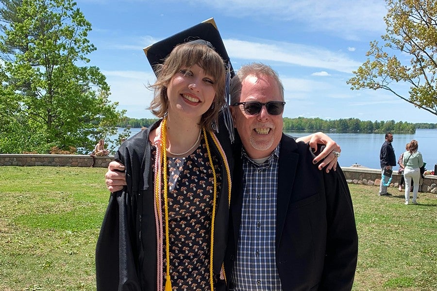A smiling student in a graduation robe stands with her arm around her arts instructor.