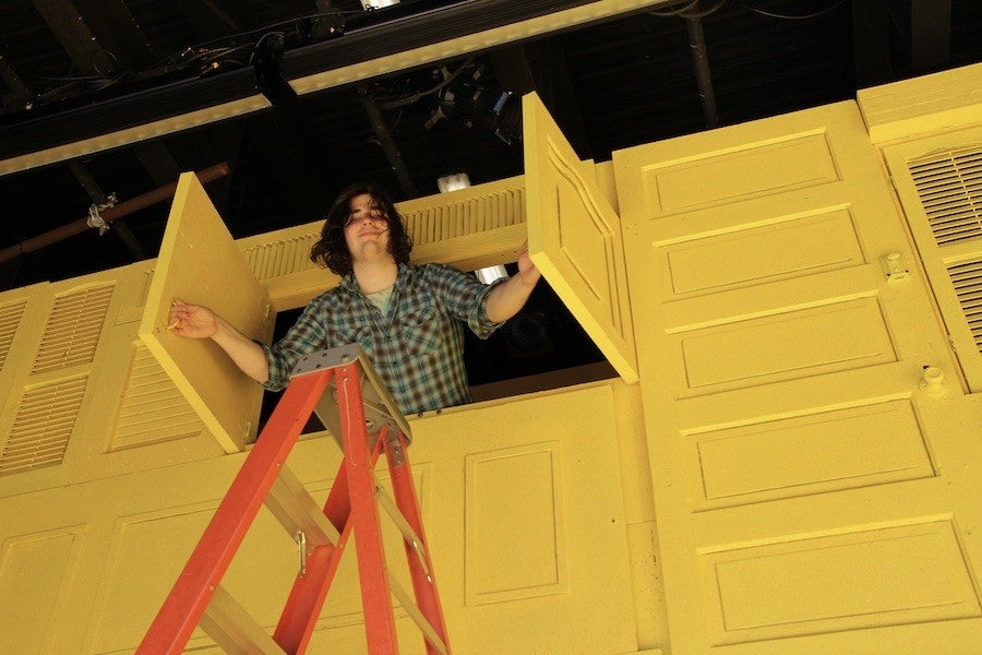 A smiling person leans out of a wall of yellow windows.