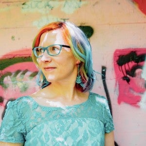 A woman with brightly colored hair stands in front of a graffiti background.