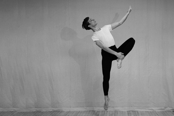  A dancer in black pants and a white shirt leaps with one leg extended downward and  the opposite foot touched to the inner thigh, extending one arm elegantly upward, captured in black and white.