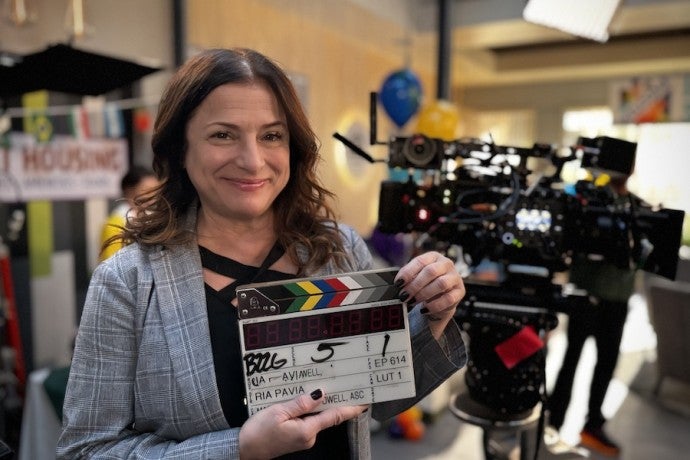 A smiling director poses with a clapperboard in front of camera equipment.