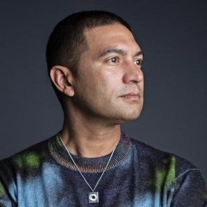 A dark-skinned man wearing a multicolored shirt and silver necklace gazes to the viewer's right.