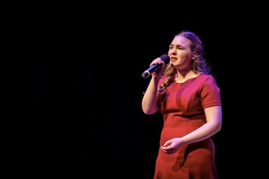 A young woman in a red dress sings soulfully into a microphone.