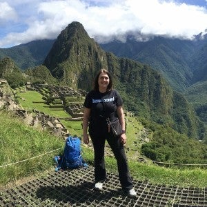 A woman dressed in black hiking clothes stands in front of a green and mountainous landscape with ancient ruins.