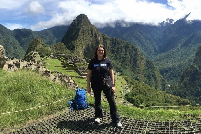 A woman dressed in black hiking clothes stands in front of a green and mountainous landscape with ancient ruins.