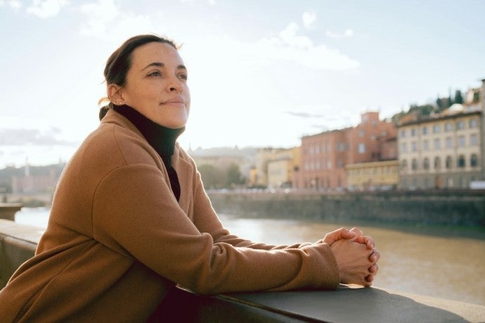 A woman dressed in a camel coat stares pensively from her perch on a bridge.