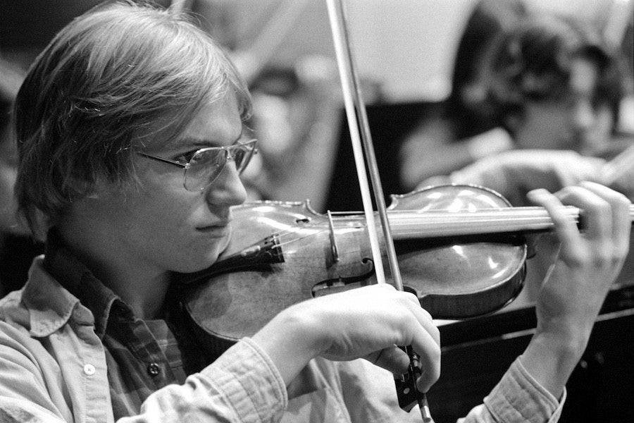 Young musician in glasses playing a violin focusedly, in a black and white photo, with another person faintly visible in the background.